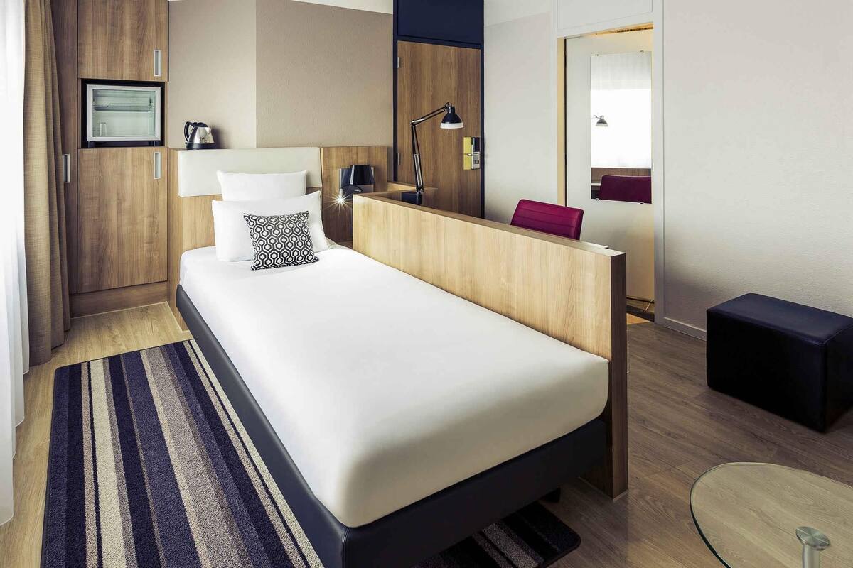 Ein modernes Hotelzimmer mit einem großen Bett, einem hölzernen Kopfteil, einem Nachttisch mit Lampe, einer kleinen Sitzecke und einem Spiegel.