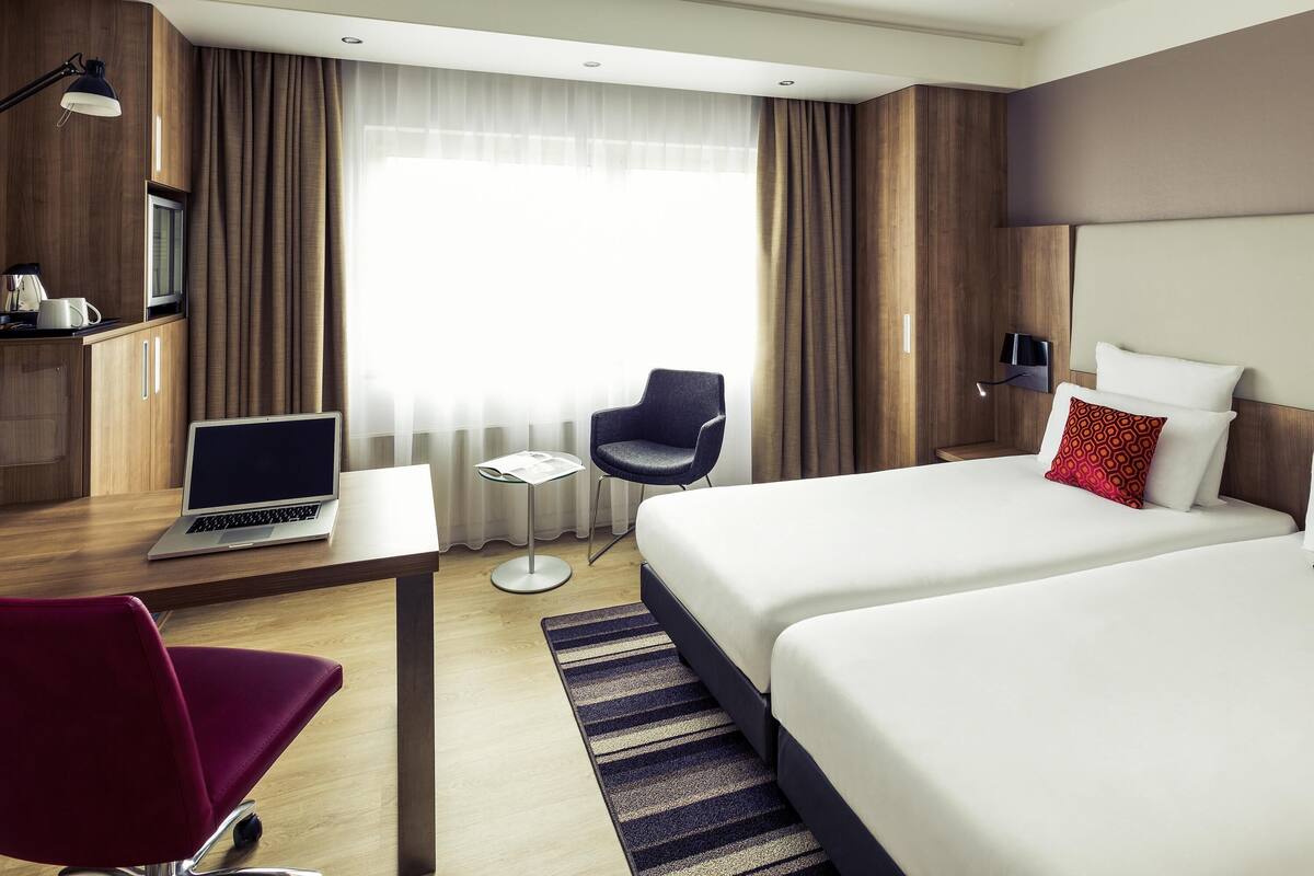 Ein Hotelzimmer mit Schreibtisch, Laptop, Stuhl, zwei Betten und einem Fernseher.