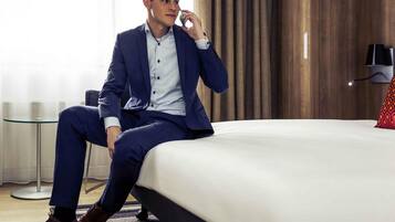 A man sitting on a bed in a hotel room with a wooden headboard, a glass table, and a red pillow.