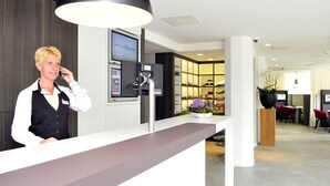Zona de recepción de hotel con diseño moderno, una mesa de recepción, una máquina de café y un área de descanso con sillas y una mesa.