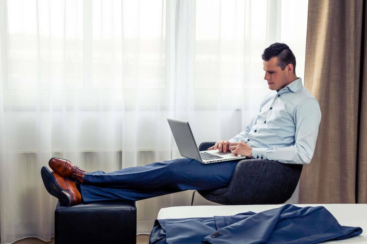 Eine Person sitzt auf einem Bett, ein Laptop auf dem Schoß, und trägt ein Hemd und eine Hose.