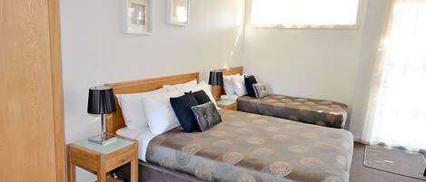 A hotel room with a double bed, a single bed, and a nightstand with a lamp. There are two framed pictures on the wall and a window with curtains.
