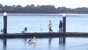 A pier with a person fishing, another person standing, and a pelican swimming in the water.