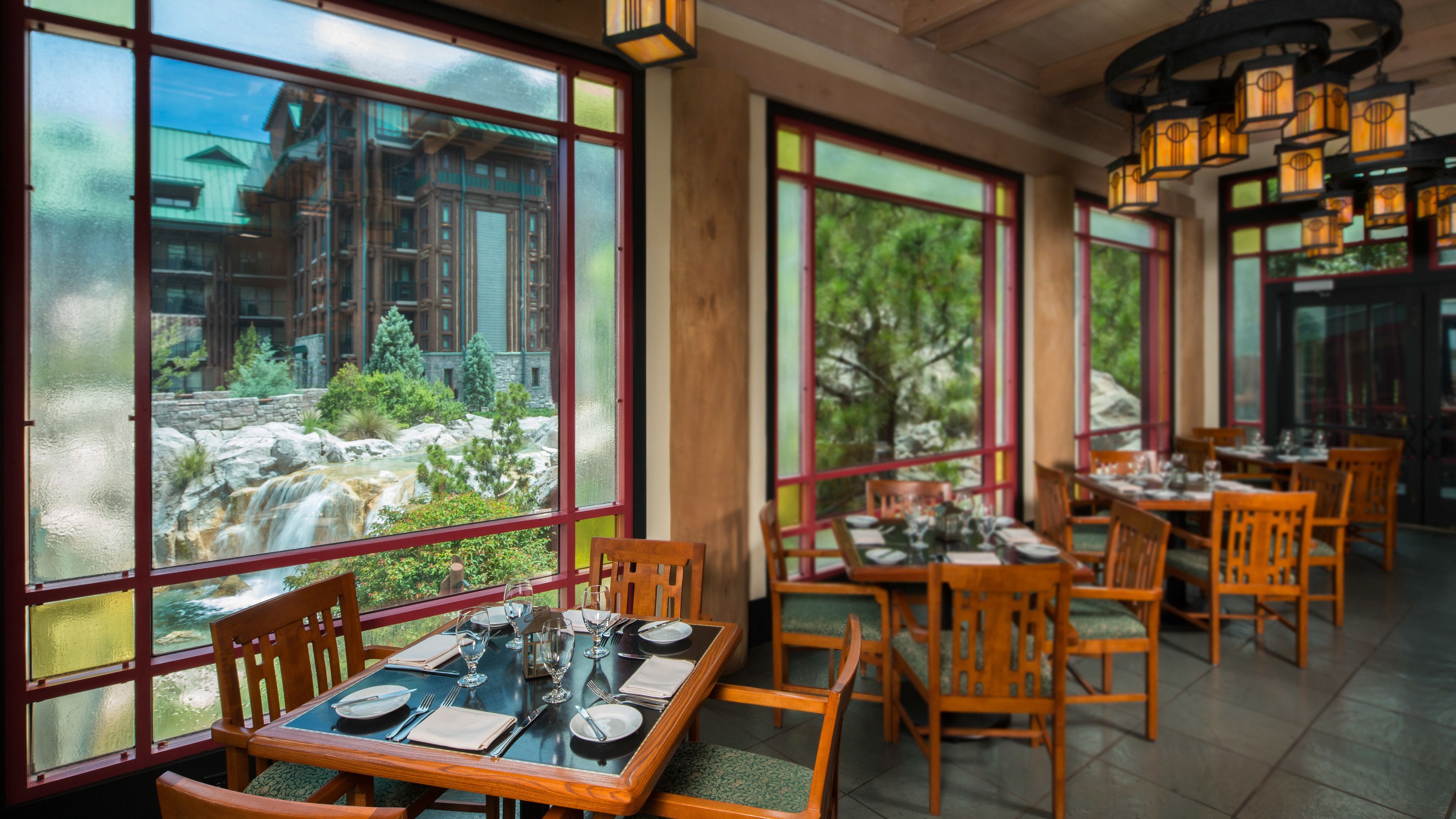 Een restaurant met houten tafels en stoelen, grote ramen met uitzicht op een waterval en groen, en hangende lantaarns.