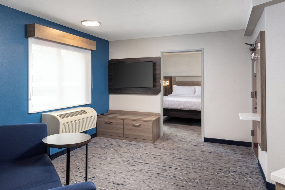 A hotel room with a large flat-screen TV mounted on the wall, a bed with white bedding, a small round table with a chair, and a built-in air conditioning unit.