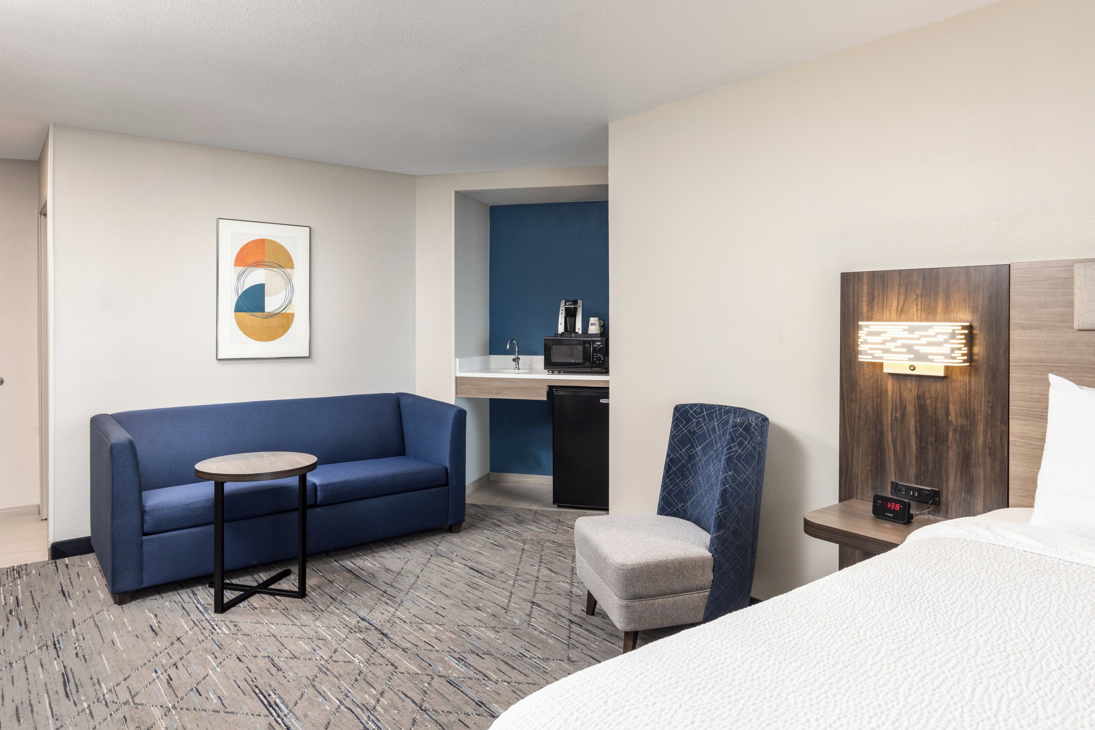 Una habitación de hotel con una cama, un sofá azul, una mesita, una silla, una mesita de noche con un reloj, un microondas y una cafetera.
