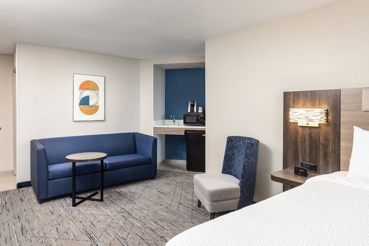 Une chambre d’hôtel avec un lit, un canapé bleu, une petite table, une chaise, une table de chevet avec une horloge, un four à micro-ondes et une machine à café.