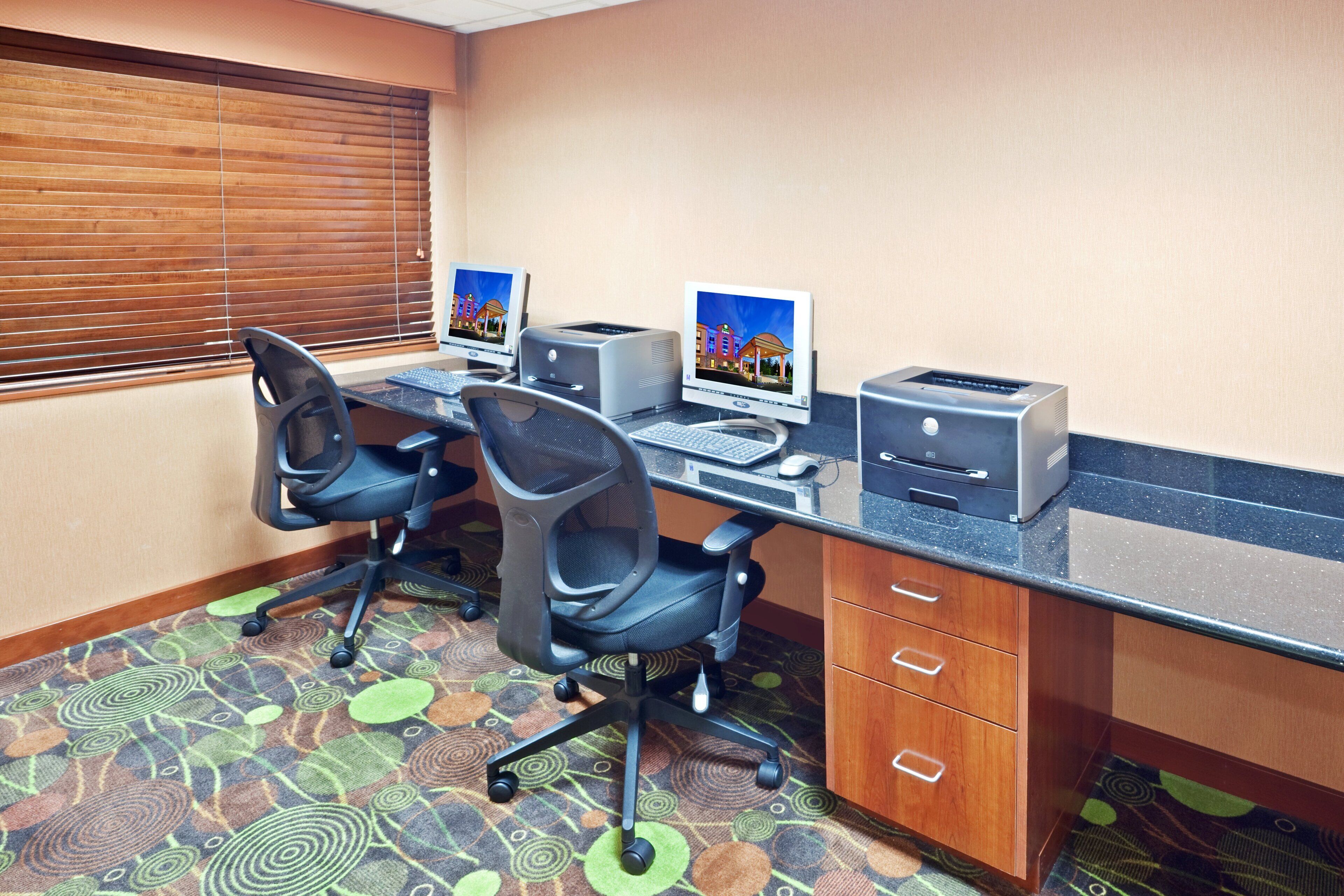 A hotel room with two desks, two chairs, a desktop computer, a printer, and a fax machine.
