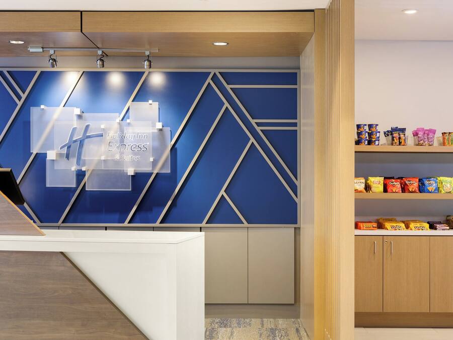 A hotel reception area with a blue geometric wall design, a clear sign for Holiday Inn Express & Suites, and a snack shelf with various packaged foods.