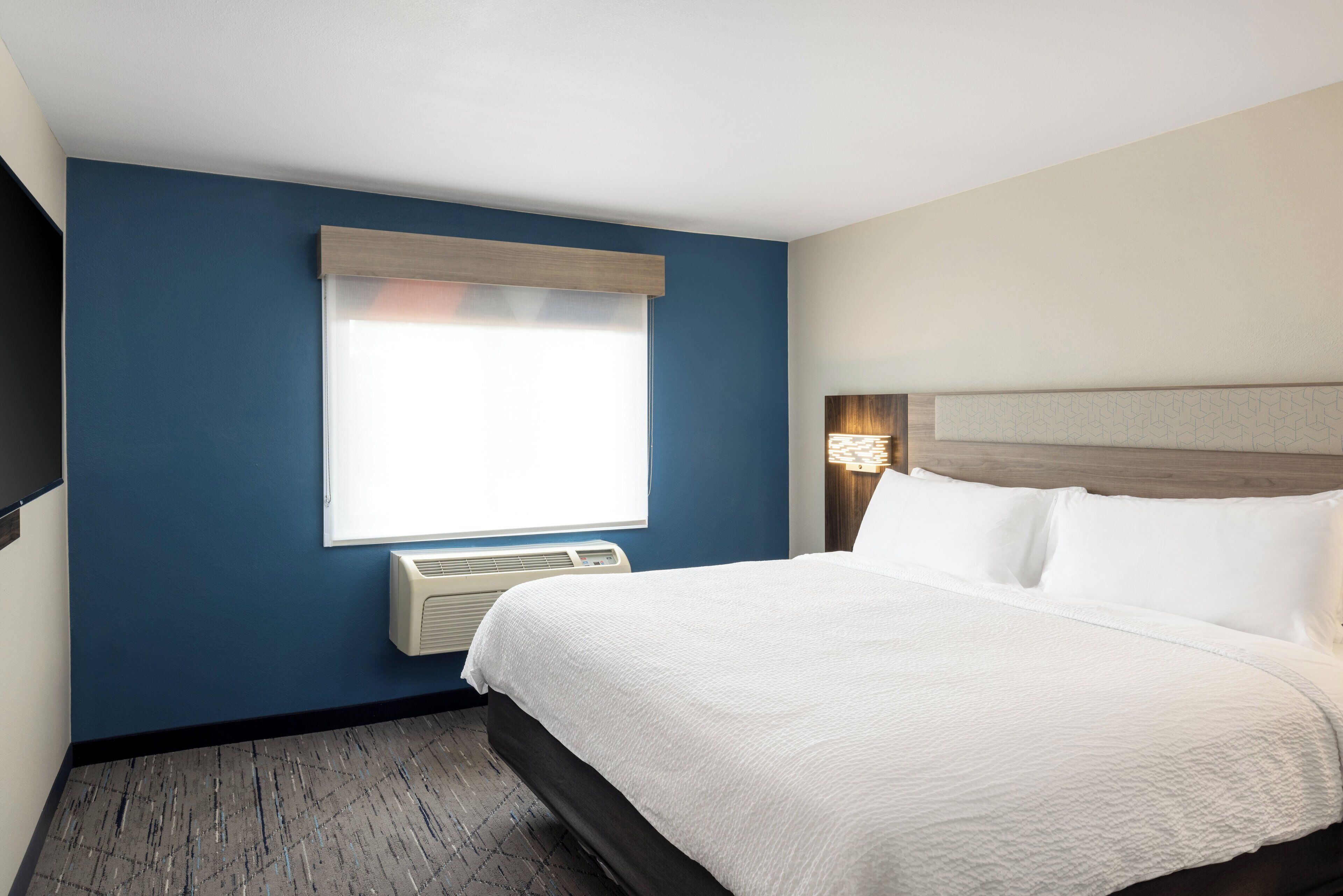 A hotel room with a large bed, a window with blinds, a wall-mounted air conditioner, and a headboard with a light fixture.