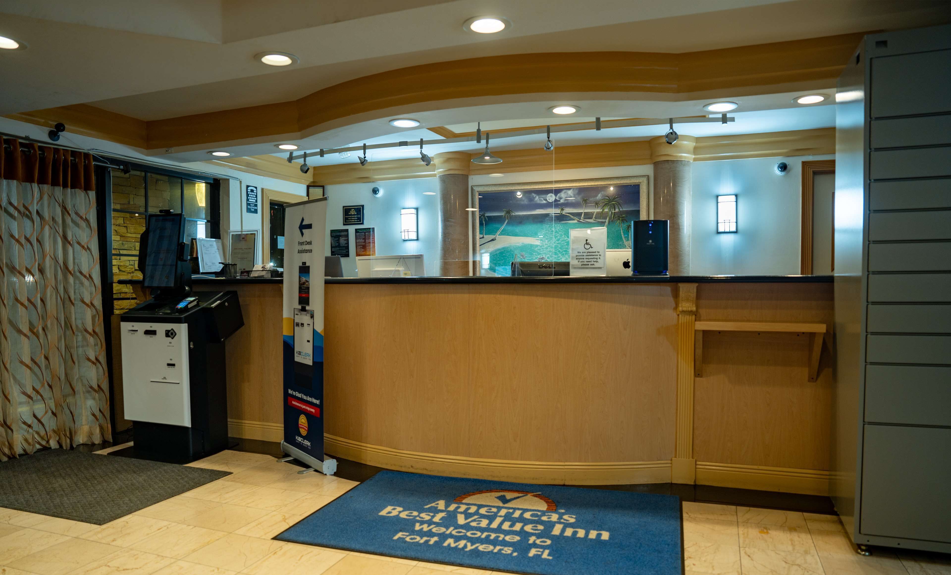 A reception area with a wooden reception desk, a computer, and a sign for front desk assistance. There is a blue mat with "Best Value Inn" and "Welcome to Fort Myers, FL" text.