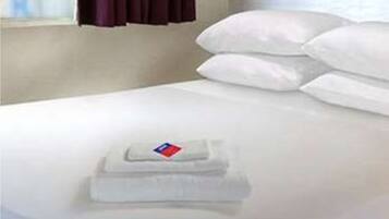 A hotel room with a bed, white bedding, a window with purple curtains, and a small table with a towel and a flag.