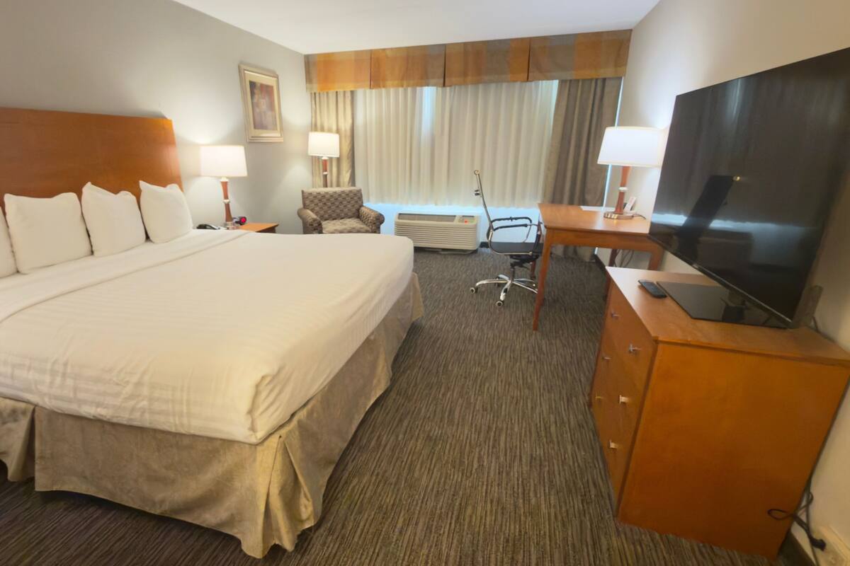 Habitación de hotel con una cama grande, un escritorio con televisor, una silla, una lámpara y una ventana con cortinas.