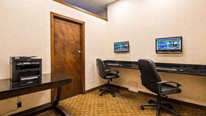 Un salón de conferencias con una puerta de madera, dos sillas de oficina negras, un escritorio negro con una computadora e impresora, y dos monitores en la pared.