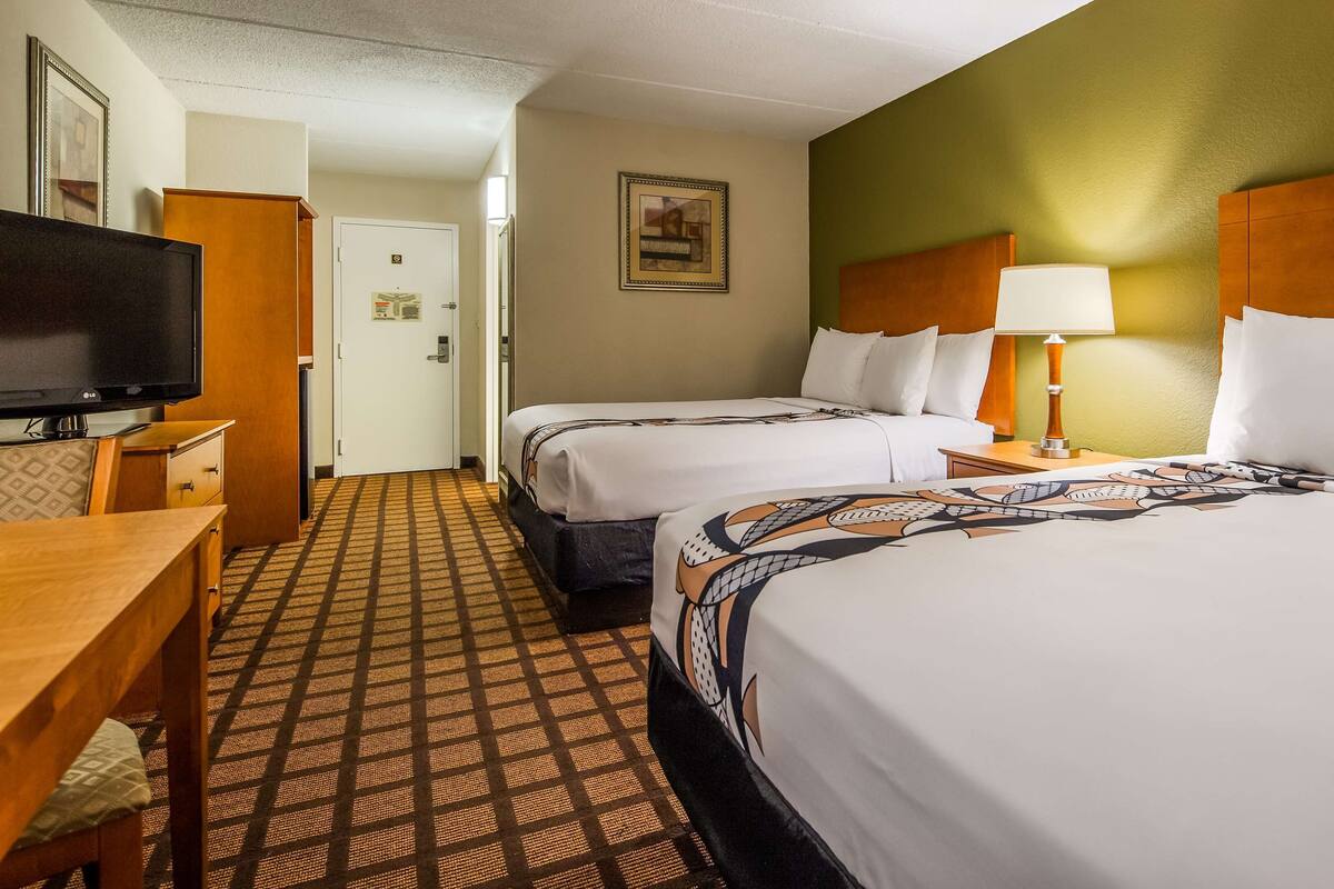 Habitación de hotel con dos camas, televisor, escritorio y alfombra estampada.