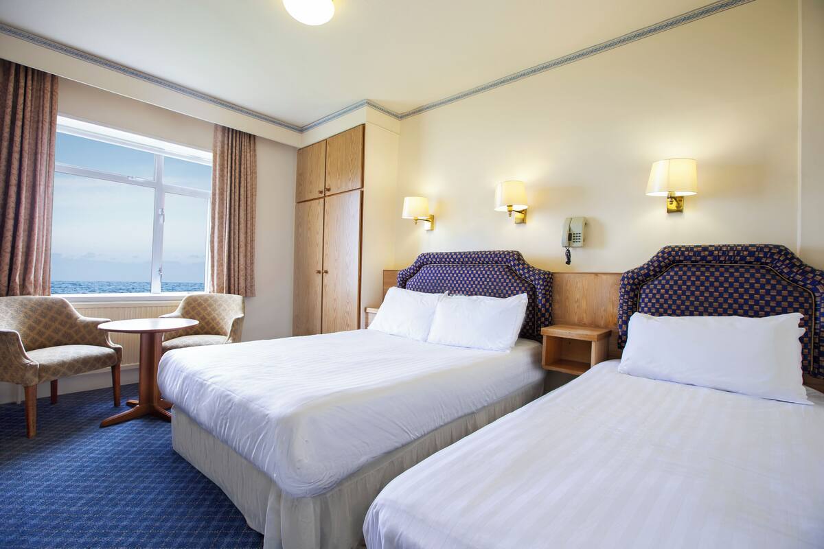 A hotel room with two beds, a small table, a chair, and a view of the sea.