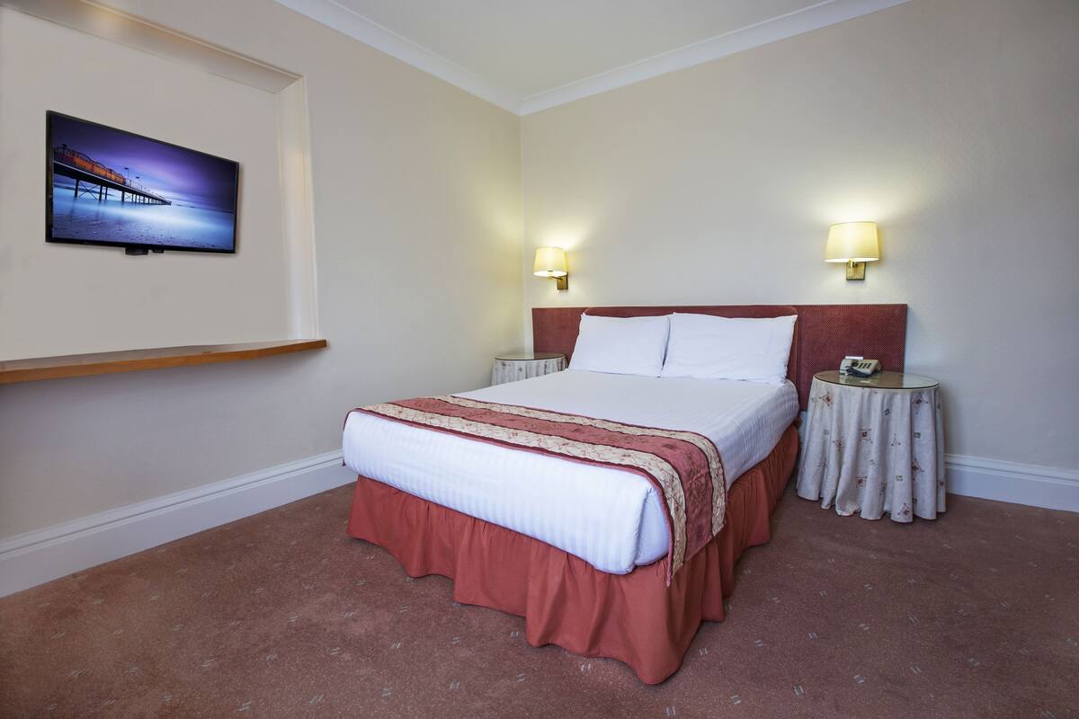 A hotel room with a bed, two bedside tables, a TV mounted on the wall, and two wall-mounted lamps.
