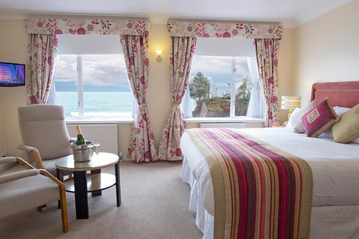 A hotel room with a bed, a chair, a small table with a bottle, and curtains with a view of the sea.