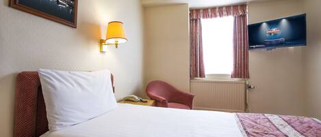 A hotel room with a bed, a chair, a TV, a lamp, and a window with curtains.