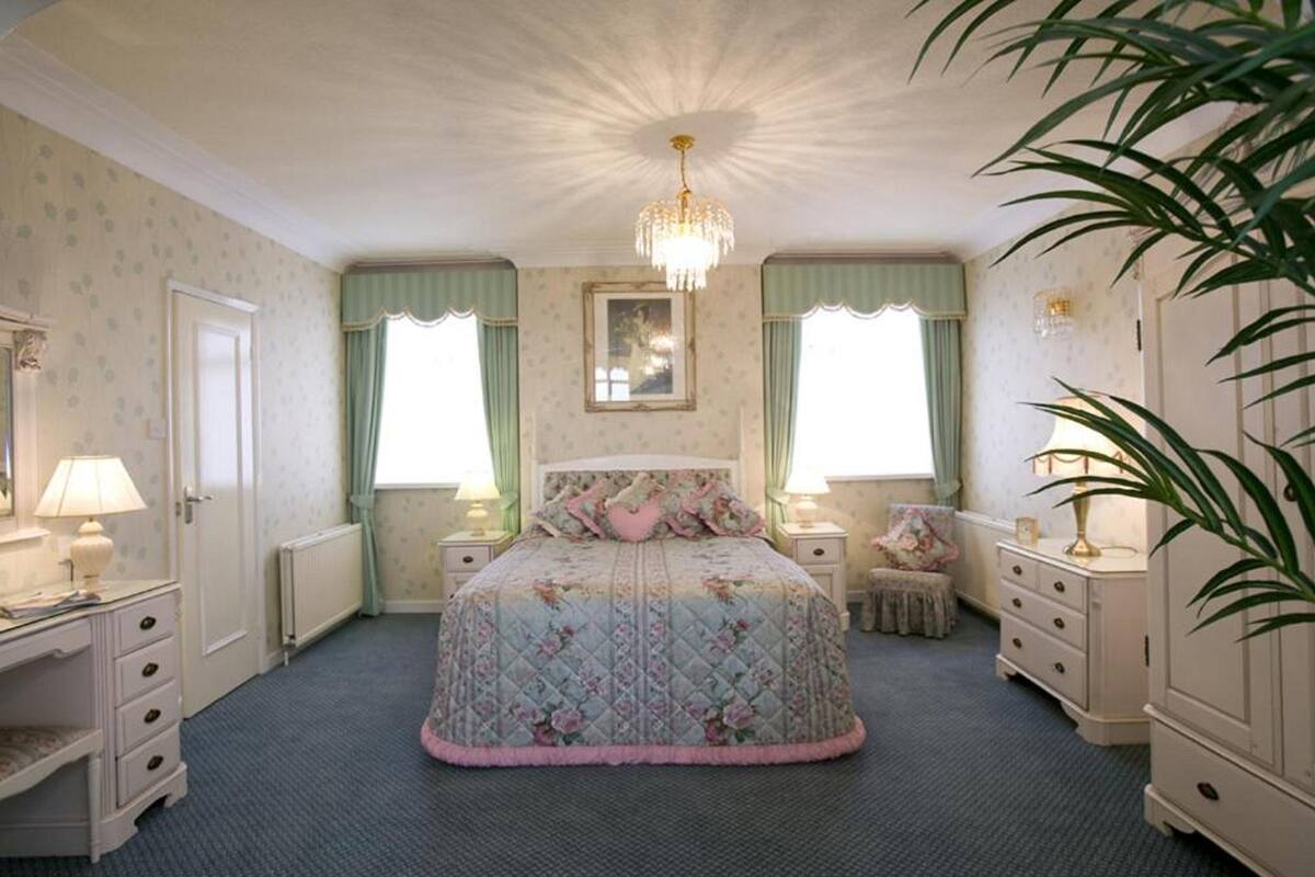 A bedroom with a large bed, two bedside tables, a dresser, a radiator, and a chandelier.