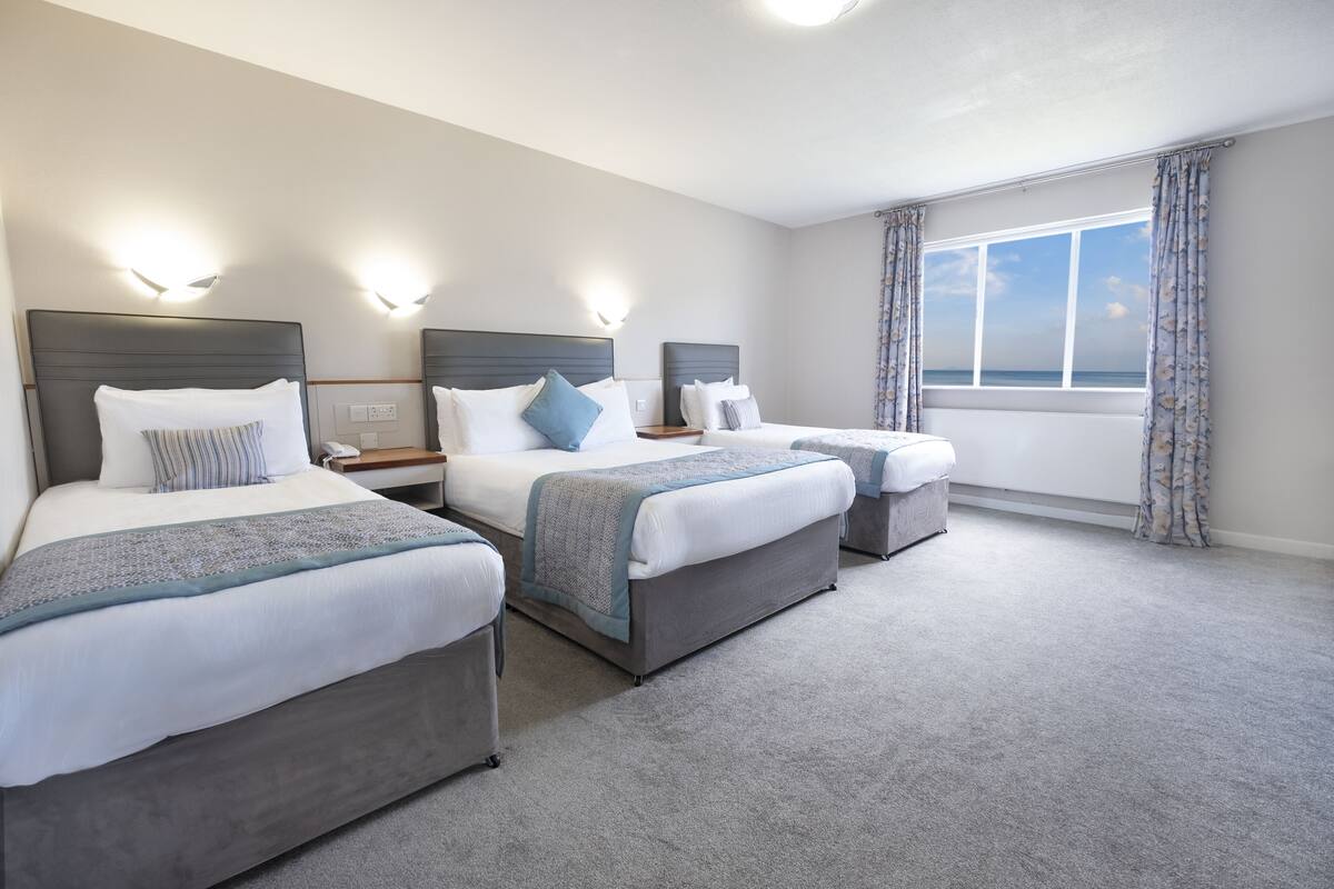 A hotel room with three beds, each with a headboard, bedside tables, and lamps. The room has a large window with curtains and a view of the sea.