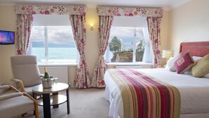 A hotel room with a bed, a chair, a small table with a bottle, and curtains with a view of the sea. - Livermead Cliff Hotel (Torquay)