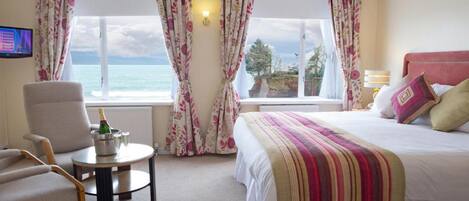 Une chambre d’hôtel avec un lit, une chaise, une petite table avec une bouteille, et des rideaux donnant sur la mer.