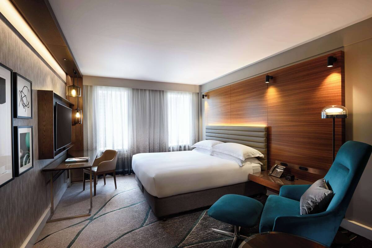 A hotel room with a large bed, a desk with a chair, a blue armchair, a TV, and a wall-mounted lamp.