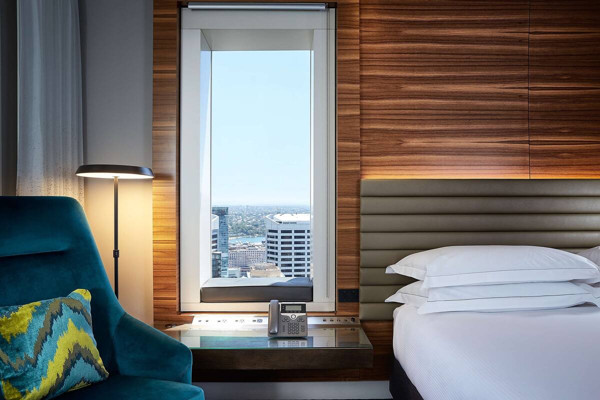 A hotel room with a view of the city, a desk with a phone, a blue armchair, and a bed with white pillows.