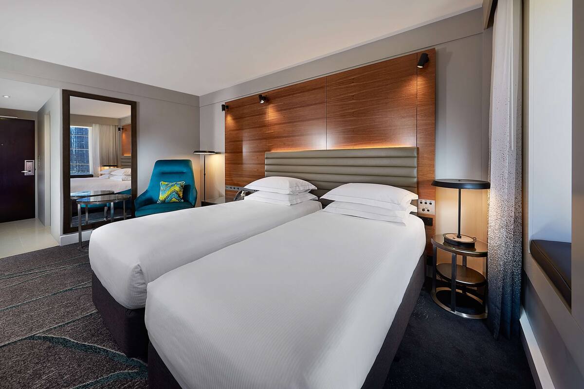A hotel room with two beds, a desk, a chair, and a nightstand with a lamp.
