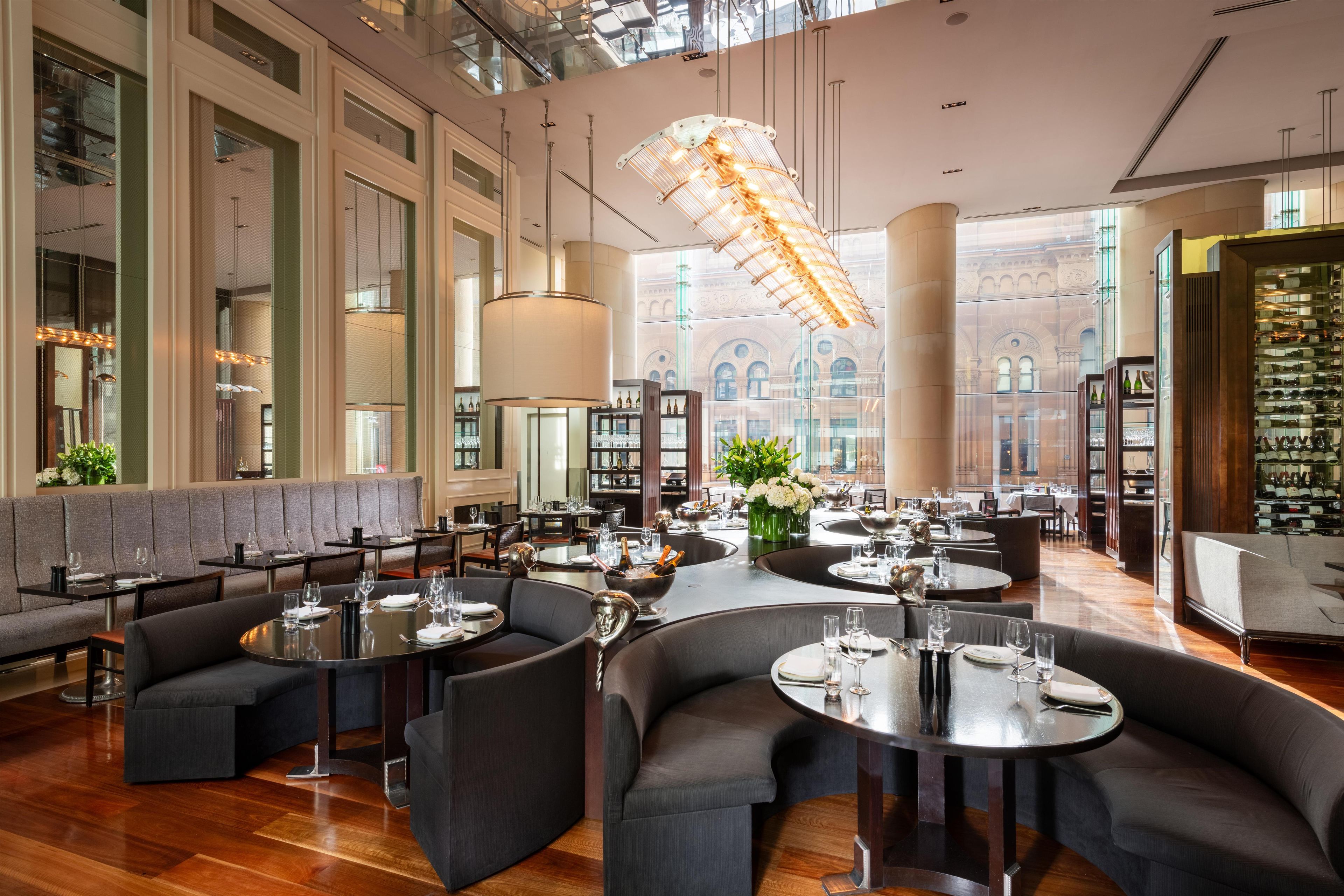 A spacious restaurant with high ceilings, large windows, and a view of historic architecture. Tables are set with wine glasses and bottles.