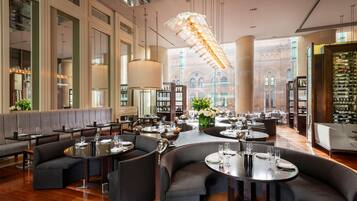 A spacious restaurant with high ceilings, large windows, and a view of historic architecture. Tables are set with wine glasses and bottles.