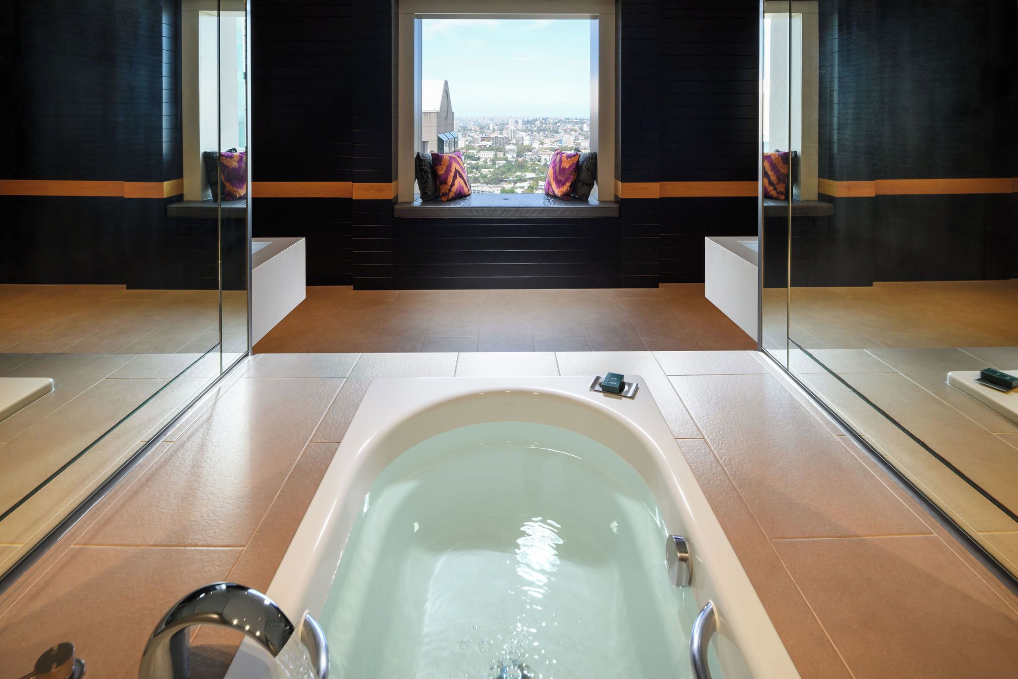 A modern bathroom with a large bathtub, a window with a city view, and a bench with cushions.