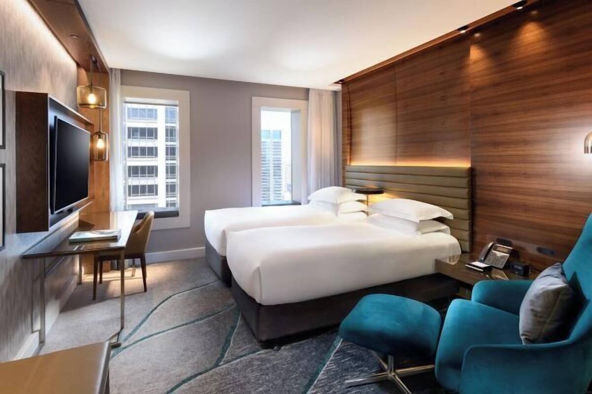 A hotel room with a bed, two bedside tables, a desk with a chair, a TV, and a window with a view of buildings.
