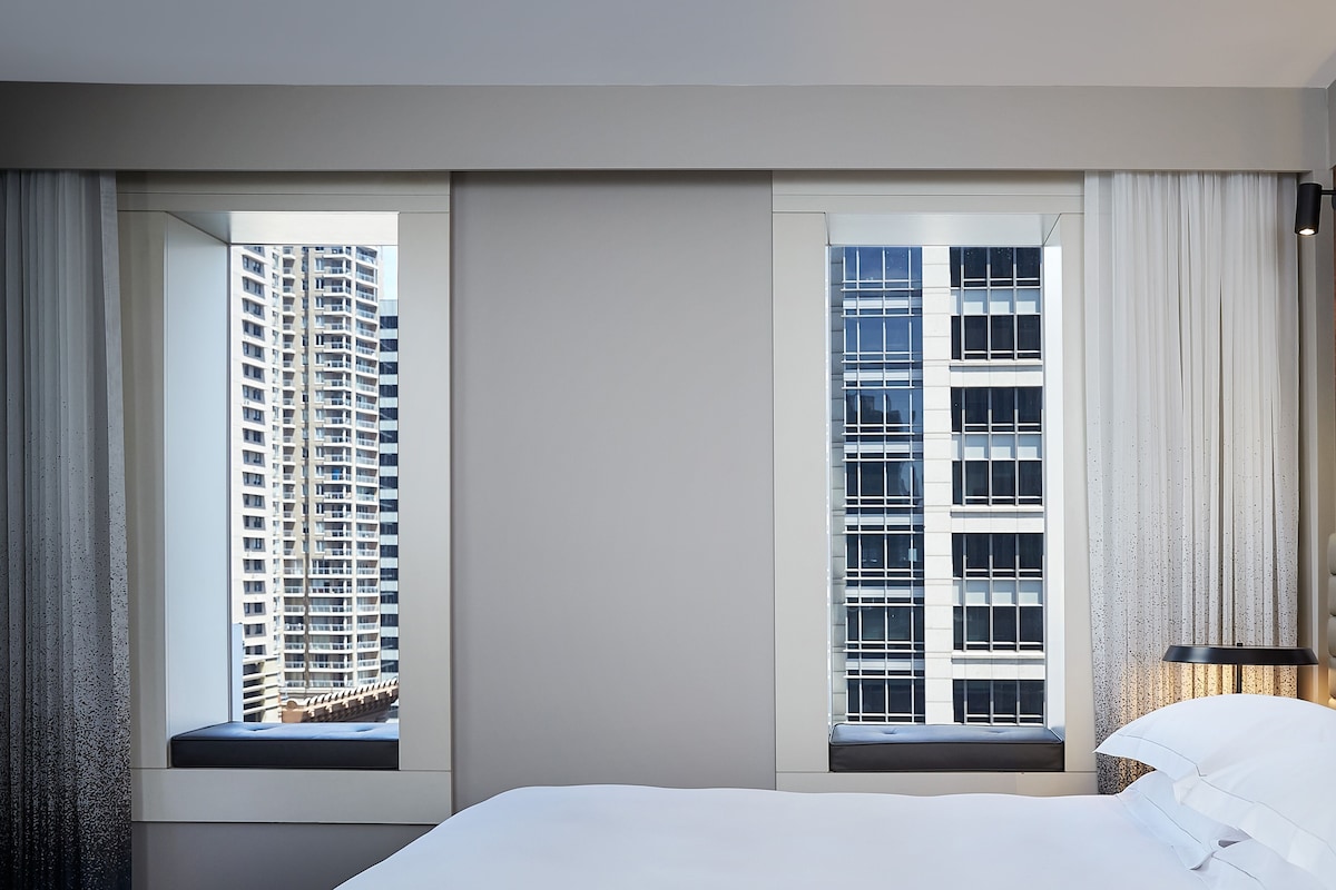 A hotel room with a large bed, two windows with views of high-rise buildings, and a bedside lamp.