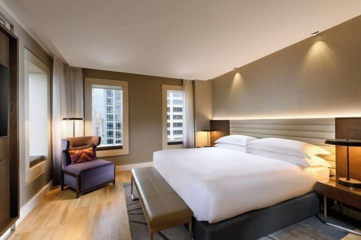 A hotel room with a large bed, a chair, a bench, two bedside lamps, and a view of buildings through the windows.
