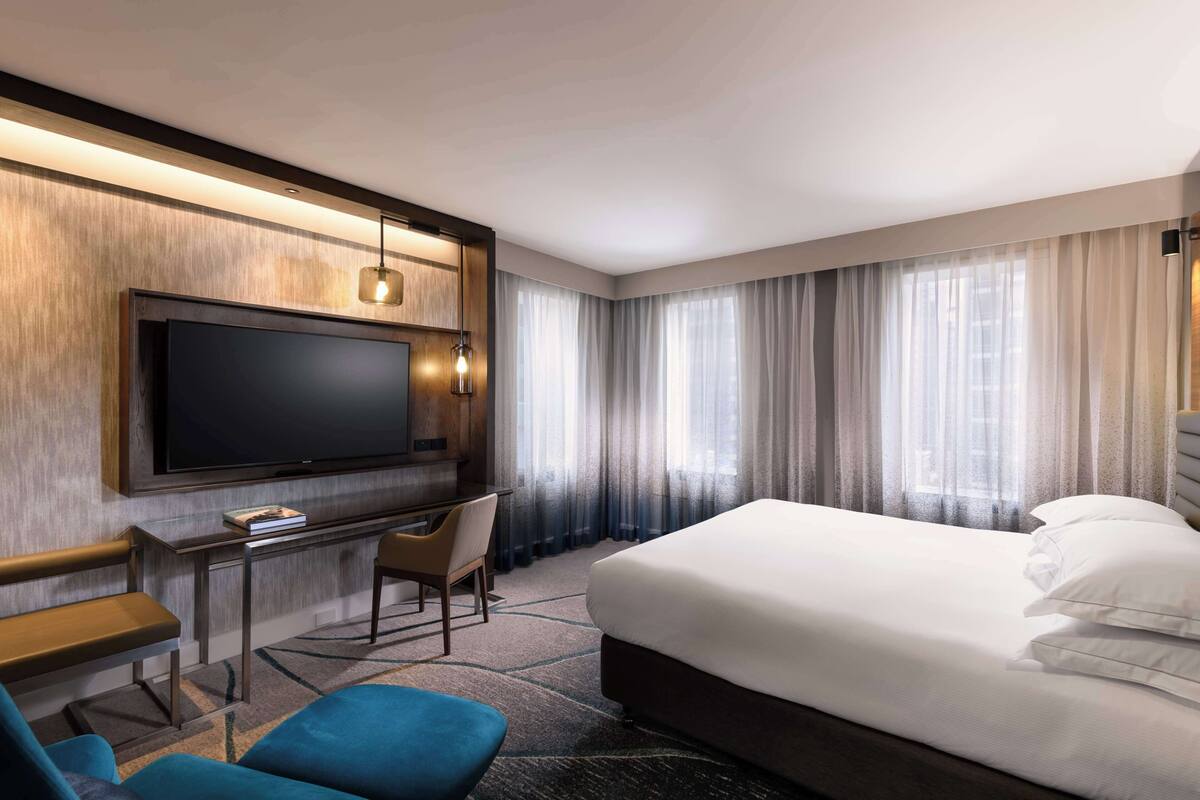 A hotel room with a large bed, a desk with a chair, a television, and a view of the city through large windows.