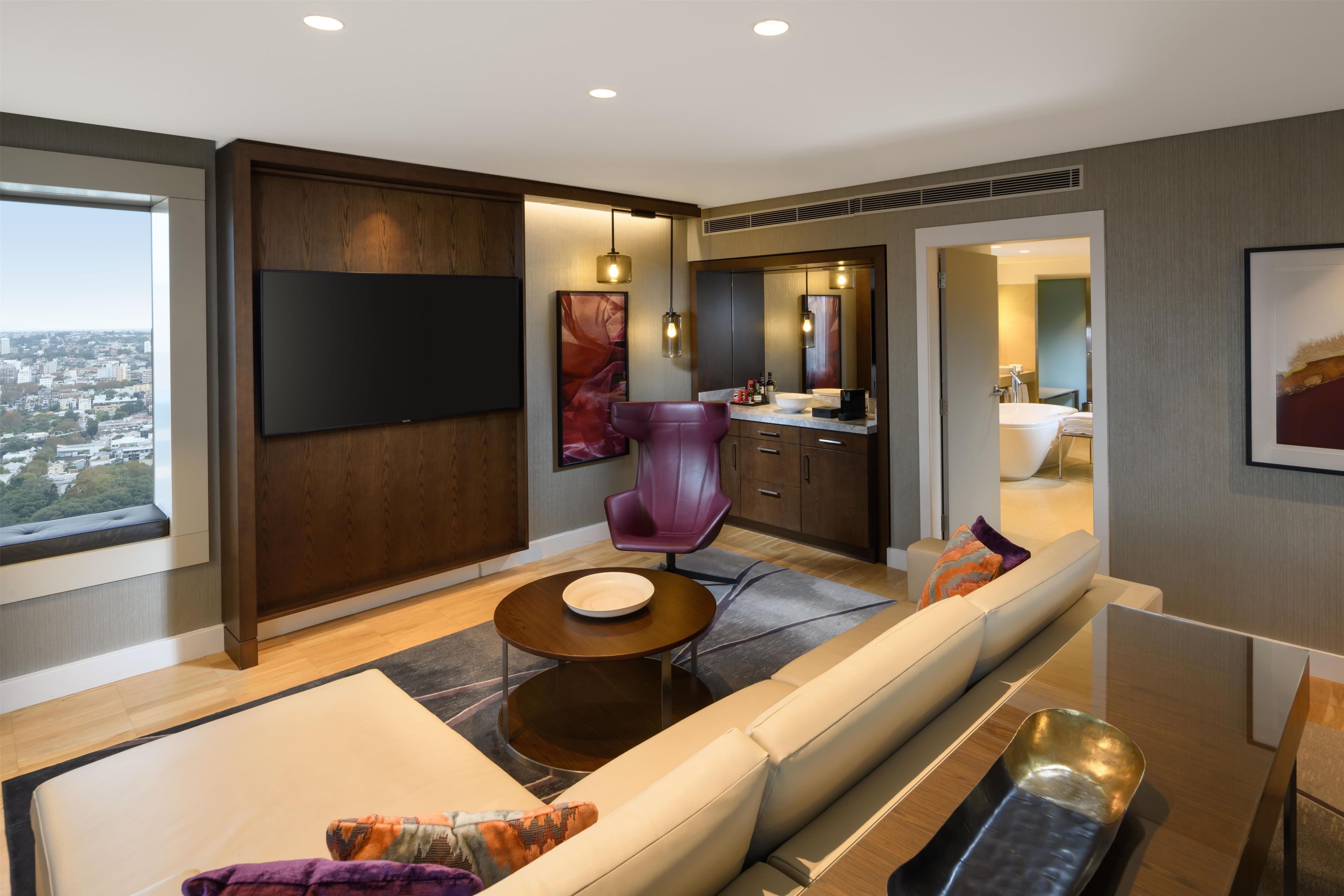 Une chambre d’hôtel moderne dotée d’une grande fenêtre, d’un canapé, d’un coin repas et d’une salle de bain visible à travers une porte ouverte.