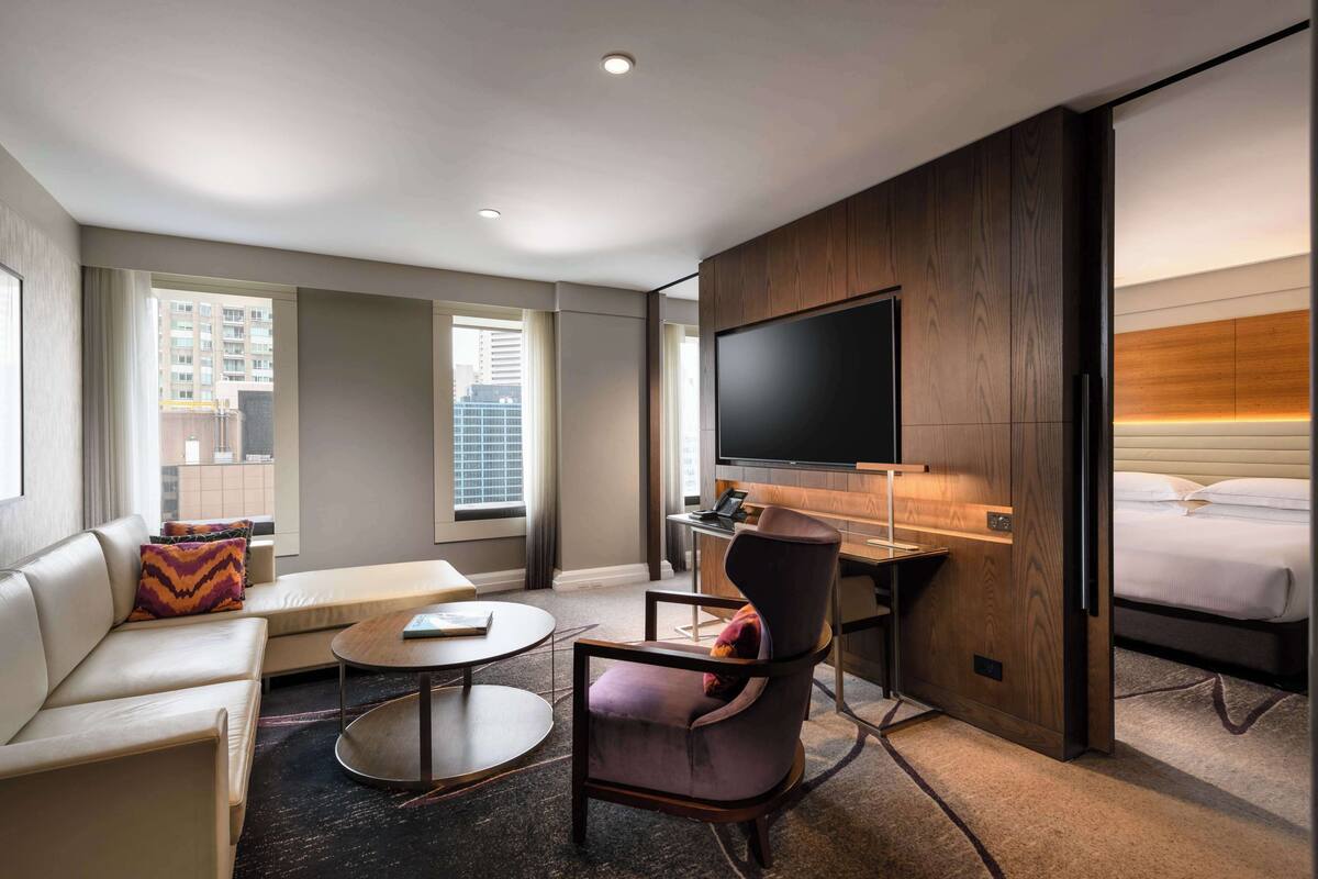A modern hotel room with a sofa, armchair, round coffee table, flat-screen TV, and a bed.