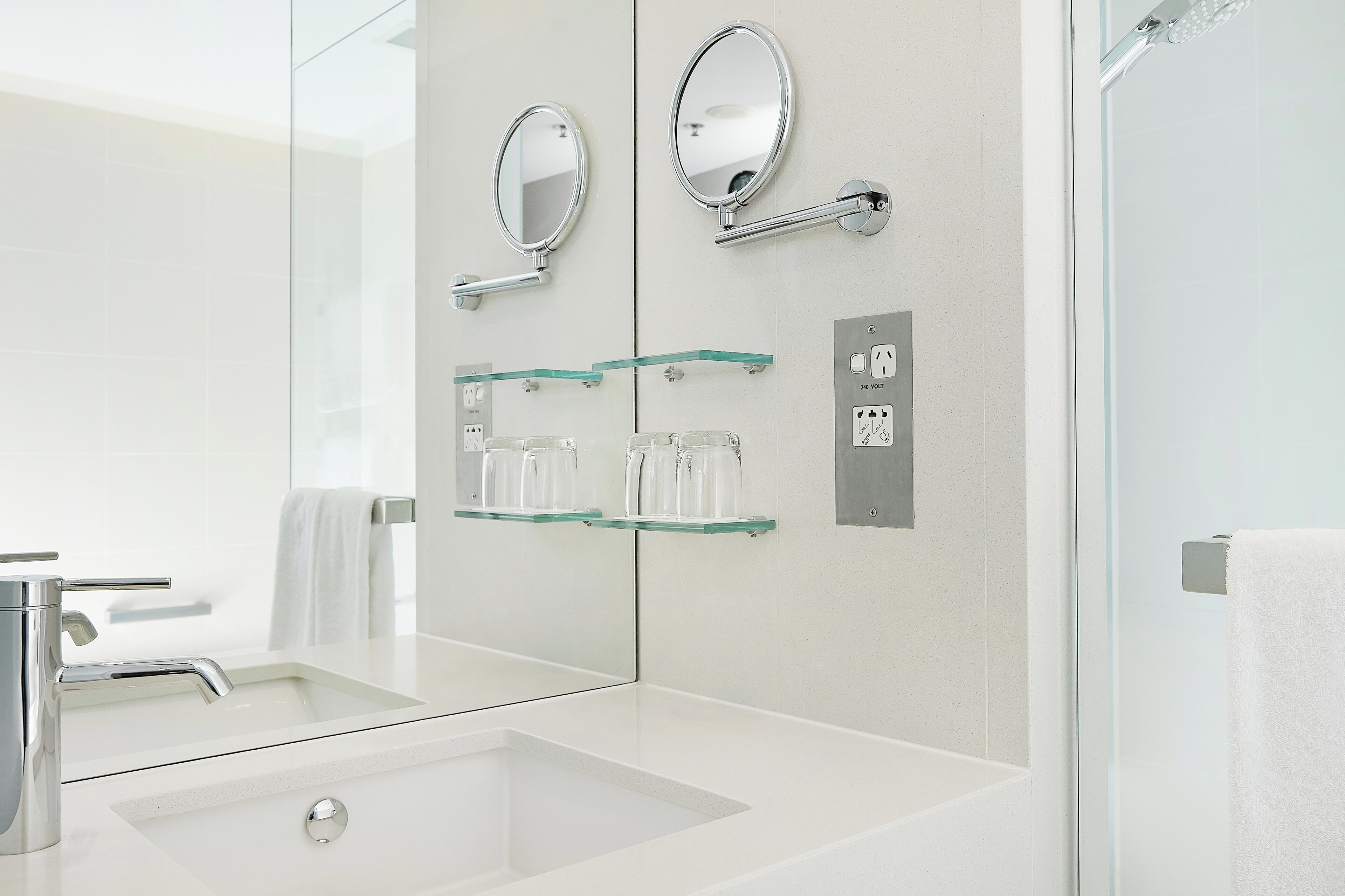 Une salle de bain moderne avec un grand miroir, une étagère en verre où sont rangés les articles de toilette et un espace douche.
