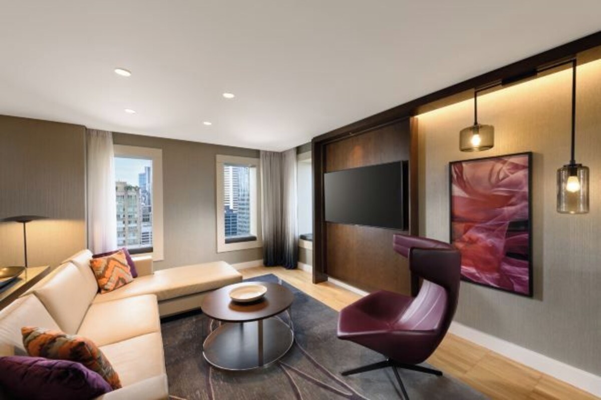 A modern hotel room with a sofa, armchair, round coffee table, and a TV mounted on the wall.