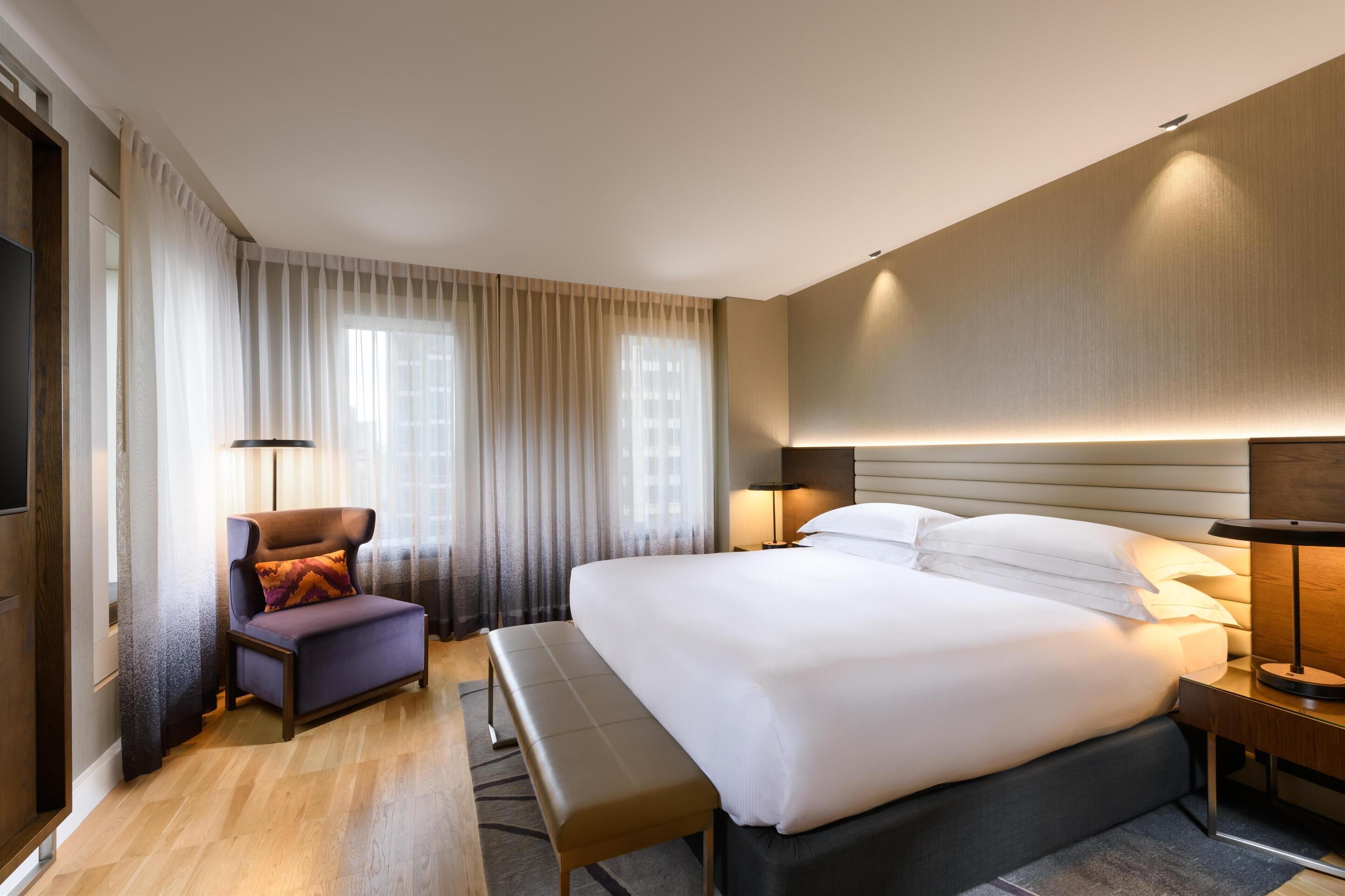 Una habitación de hotel moderna con una cama grande, un área de descanso con una silla y un banco, y una mesita de noche con lámpara.