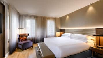 Una habitación de hotel moderna con una cama grande, un área de descanso con una silla y un banco, y una mesita de noche con lámpara.