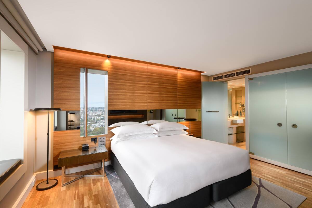 A modern bedroom with a large bed, a desk, a standing lamp, and a view of the city.
