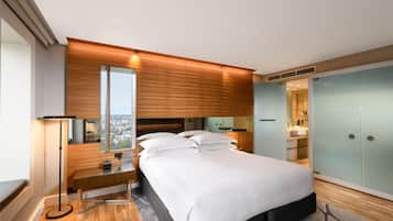 A modern bedroom with a large bed, a desk, a standing lamp, and a view of the city.
