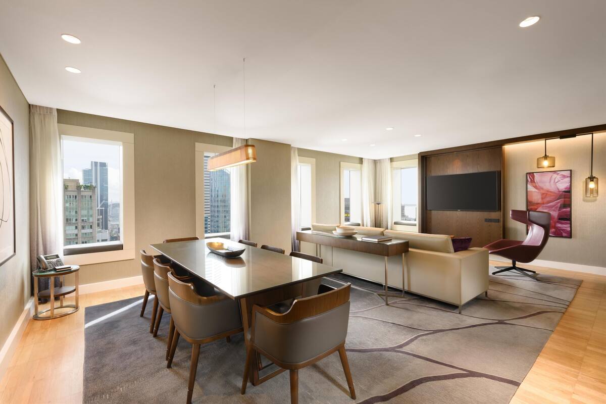 A modern hotel room with a dining area, a sofa, a TV, and a city view.