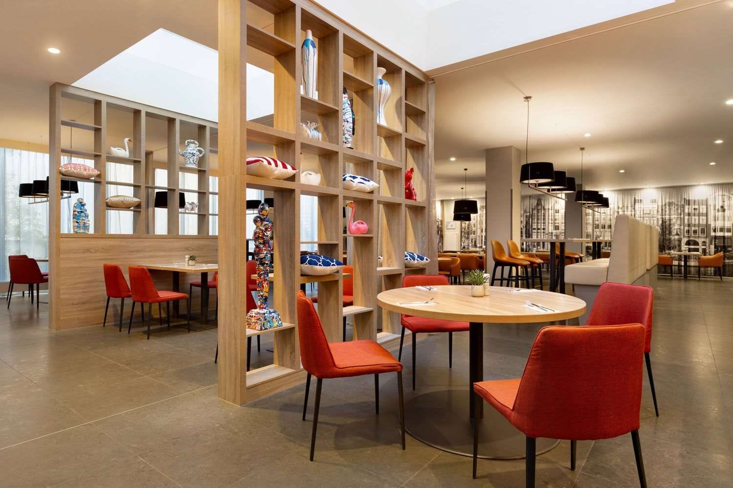 A modern interior with a round table and red chairs, wooden shelving with decorative items, and a bright ceiling.
