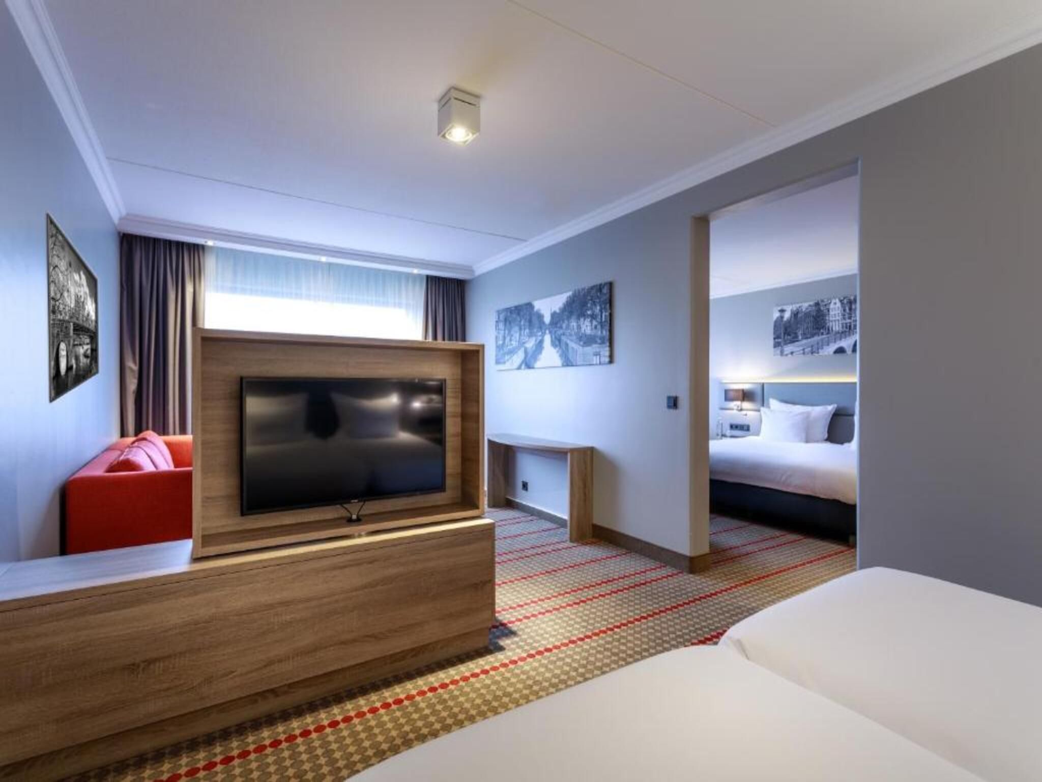 A hotel room with a large flat-screen TV, a red sofa, a bed, and a patterned carpet.