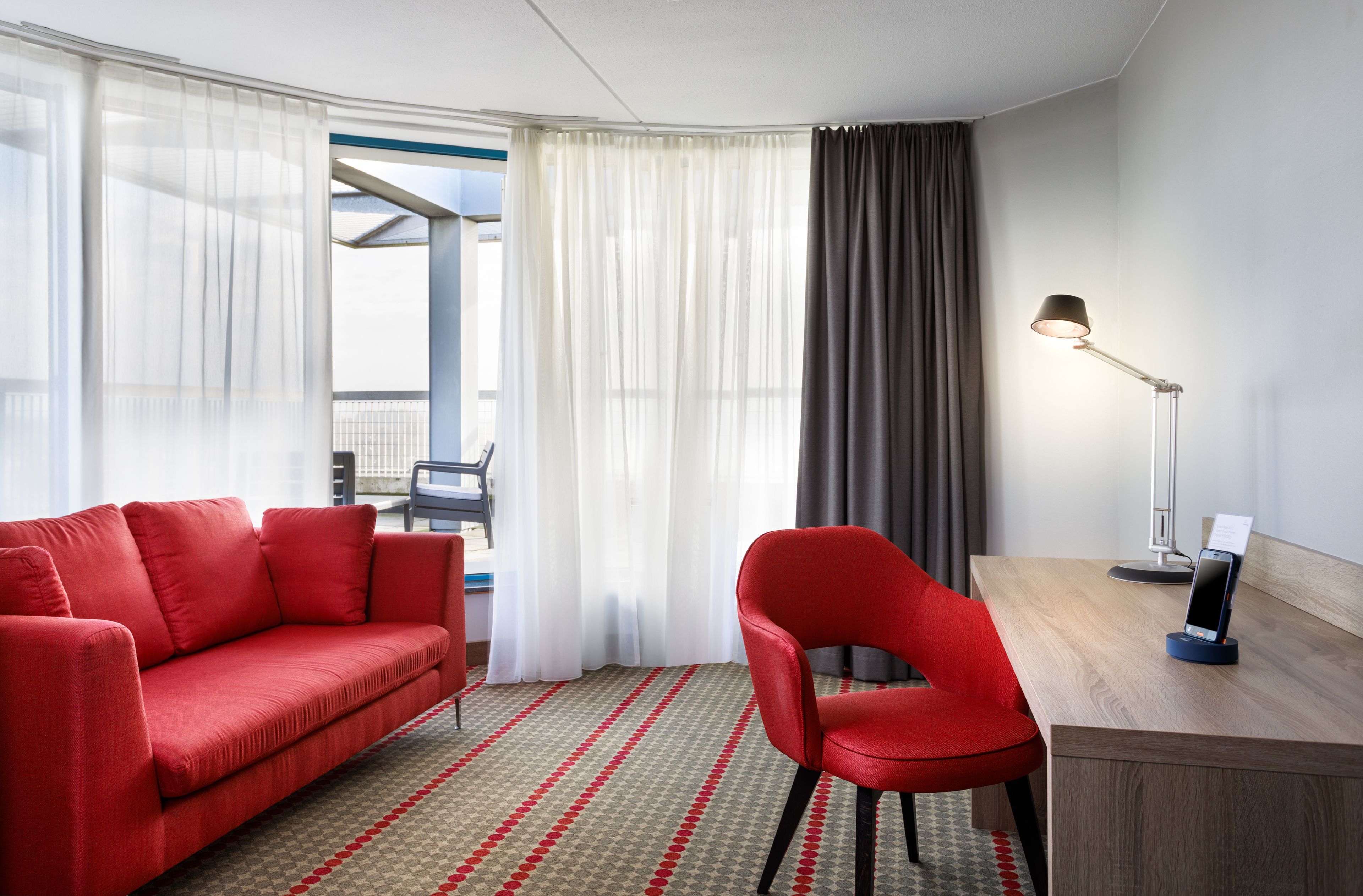A modern hotel room with a red sofa, a red chair, a desk with a lamp, and a large window with curtains.
