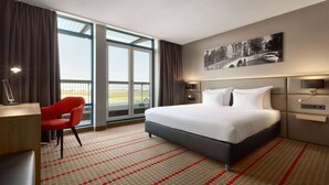 Habitación de hotel con una cama grande, un escritorio, una silla roja y un ventanal con vistas a un puente.